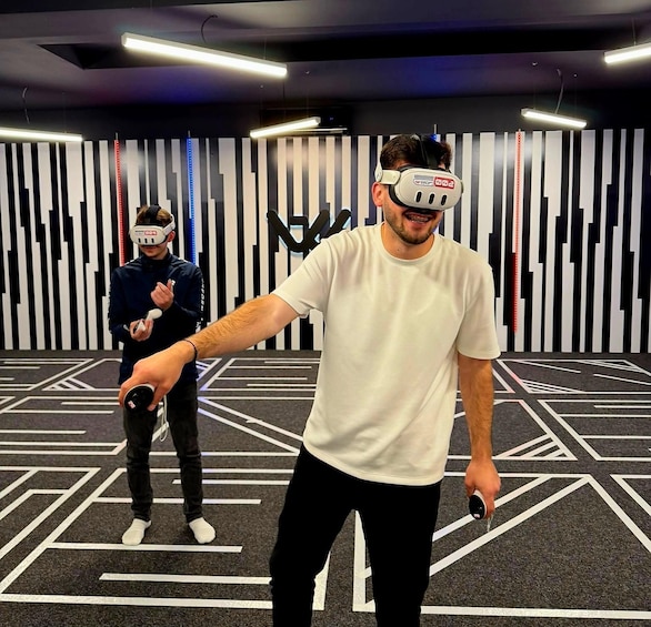 Two people in a VR gaming setup with a geometric patterned floor and a wall with vertical stripes. 3