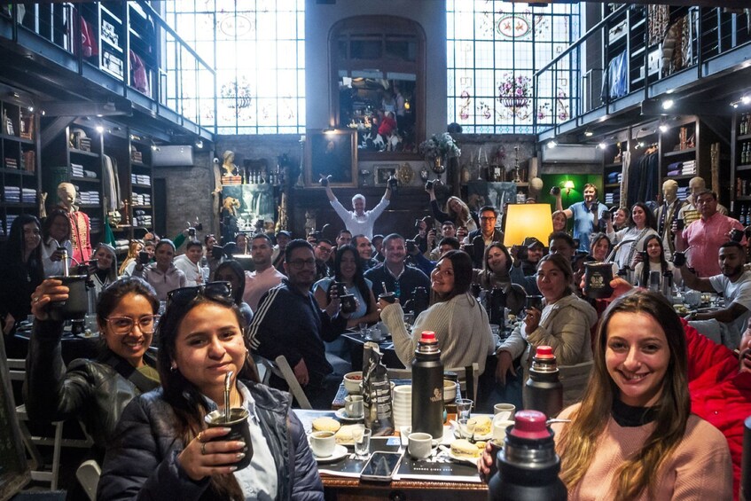 A crowded indoor venue with people seated at tables, some holding drinks, and a person in the center raising their arms. 2