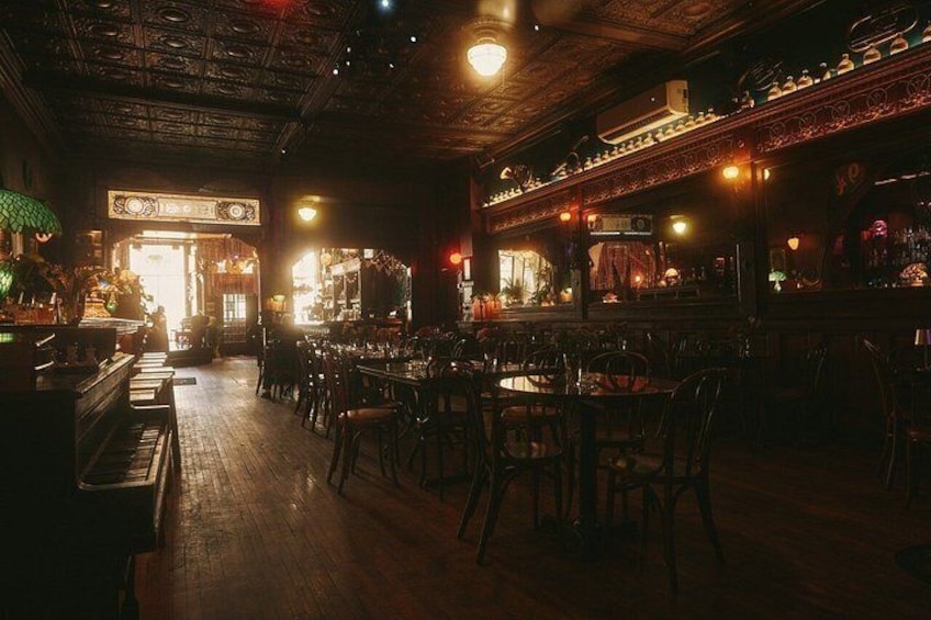 A dimly lit bar with wooden floors and tables, a bar counter with bottles, and a sign above the entrance. 3