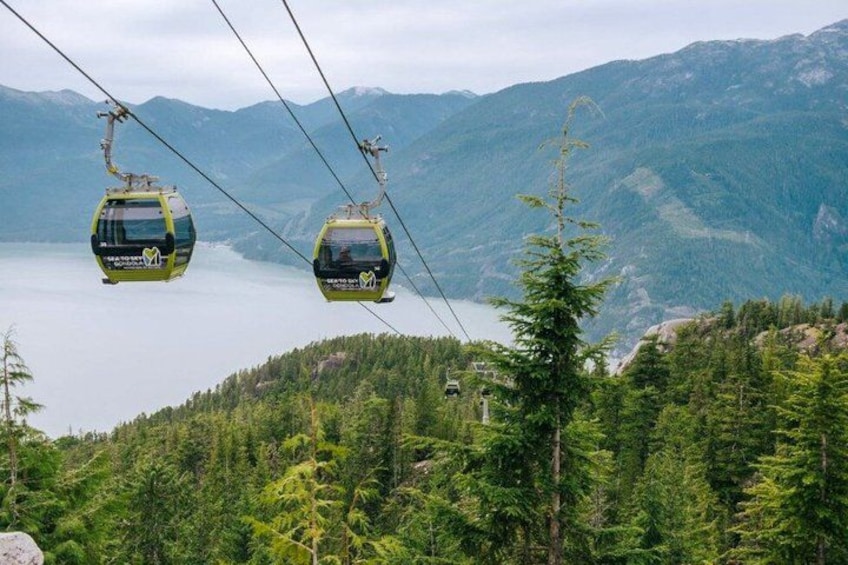 Zwei Gondelbahnen in einer Berglandschaft, im Hintergrund ein See. 1
