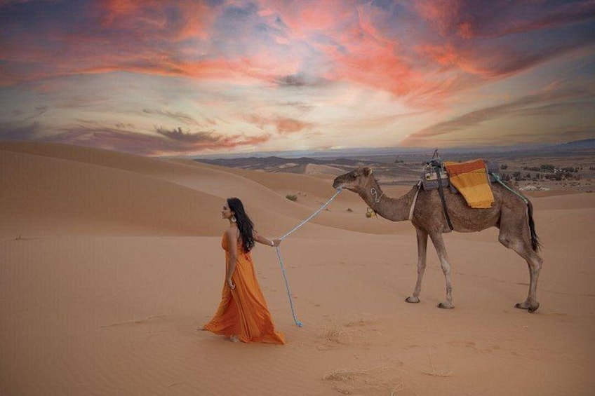 A person in an orange dress walking a camel in a desert with sand dunes and a sunset sky. 1