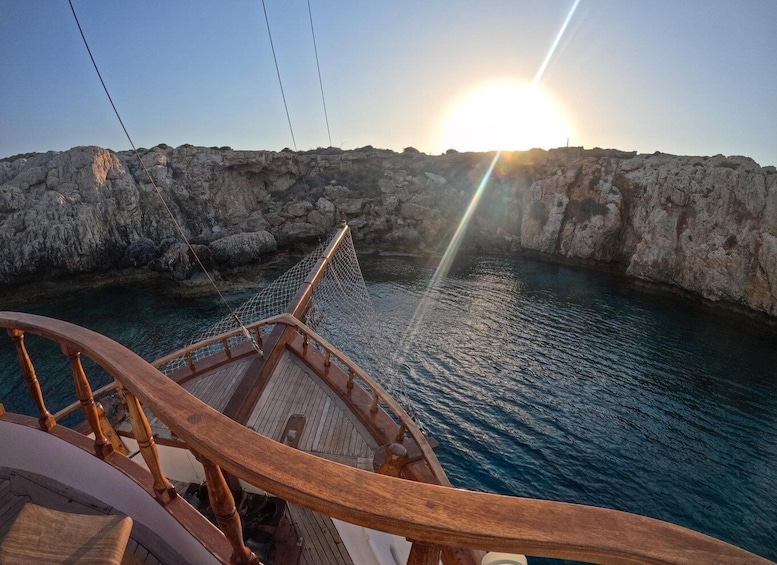 A wooden boat deck with a rope net bridge over calm water, with rocky cliffs in the background and the sun setting. 5