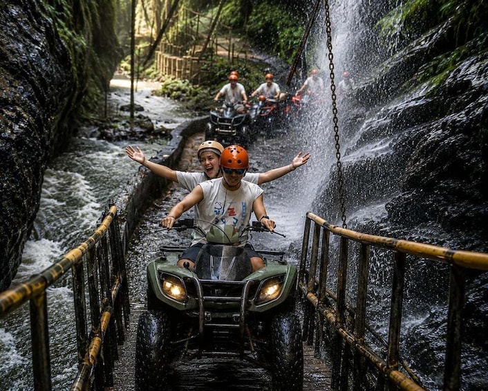 People on ATVs navigating a water-filled bridge in a forested area. 1