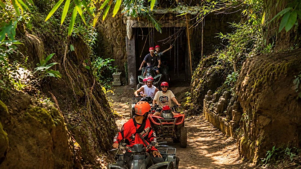 People riding ATVs on a narrow dirt path surrounded by dense vegetation and stone structures. 2