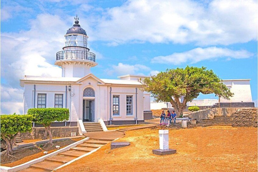 Un edificio blanco con una torre de faro, un árbol y un área pavimentada con escalones que conducen a la entrada. 4