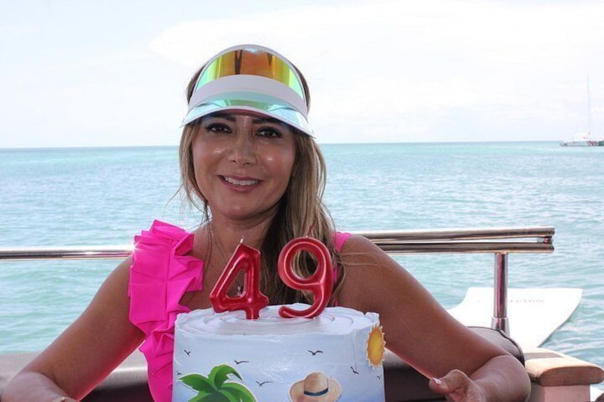 Una persona sosteniendo un pastel de cumpleaños con el número 49, vestida con un atuendo colorido y una visera, con un cuerpo de agua y un bote al fondo. 4