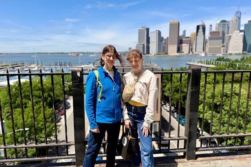 Two people standing on a bridge with a city skyline and river in the background. 4