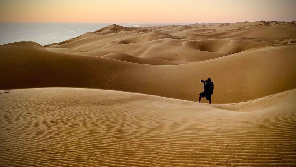 A person with a camera in a desert landscape with sand dunes and a sunset. 1