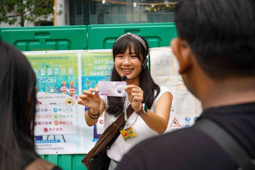 A person holding a card with text and graphics, standing in front of a green barrier with posters. 2