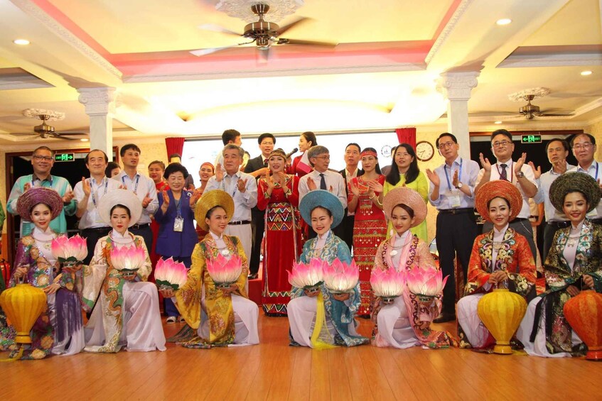 A group of people in traditional attire holding lotus-shaped lanterns, seated on a wooden floor in a room with a ceiling fan and columns. 3