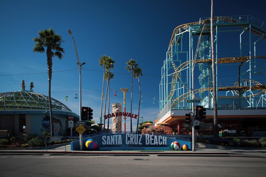 A street sign for Santa Cruz Beach Boardwalk, palm trees, and a roller coaster structure. 1