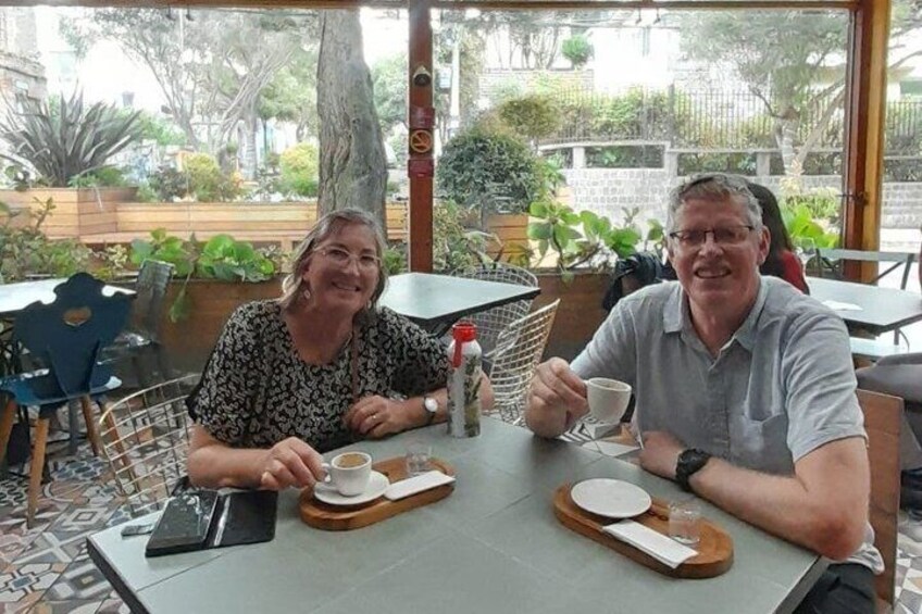 Two people seated at an outdoor cafe with a table set for two, surrounded by greenery. 5