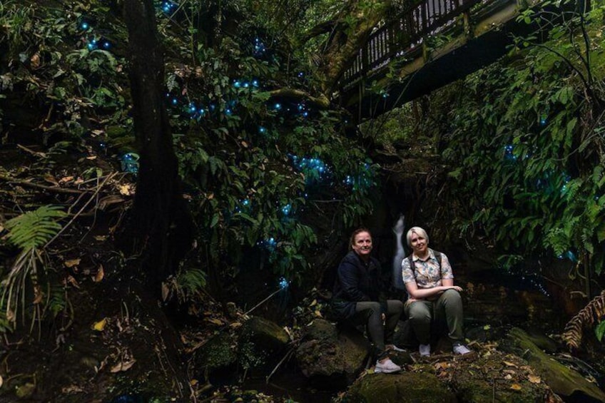 Two people sitting on a moss-covered rock in a forested area with a bridge overhead. 1