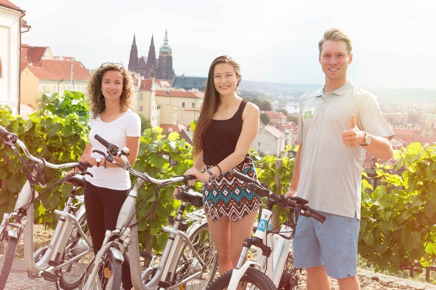 Three people standing with bicycles in a vineyard with a cityscape in the background. 4