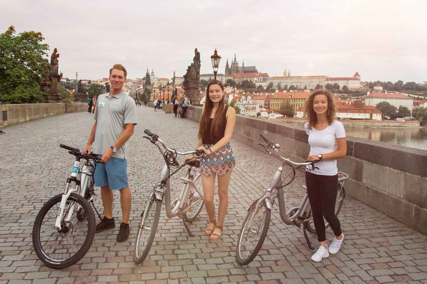 Three people standing on a bridge with bicycles, overlooking a cityscape with historic buildings and statues. 1