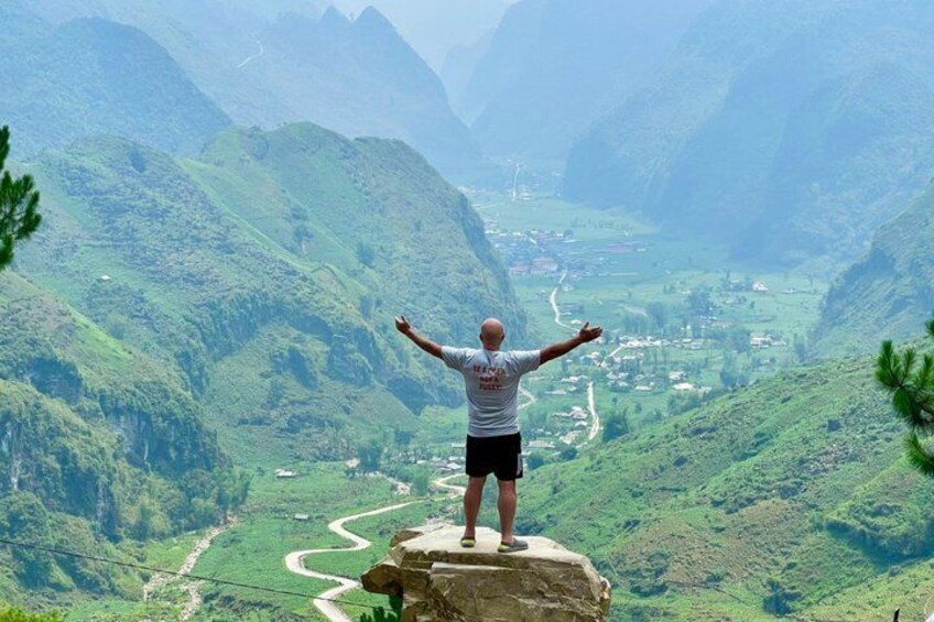 A person standing on a rock with arms outstretched, overlooking a valley with a winding road and green hills. 1