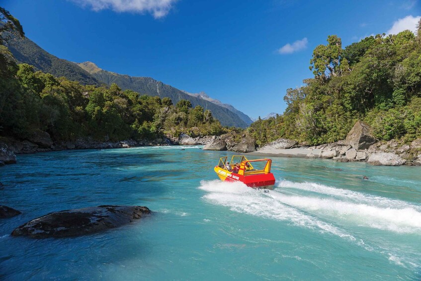 A red boat with passengers on a turquoise river surrounded by lush greenery and mountains. 1