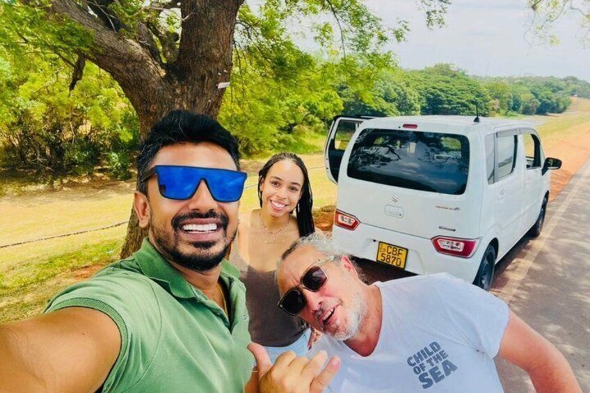 Three people taking a selfie with a white car parked on a road surrounded by greenery. 5