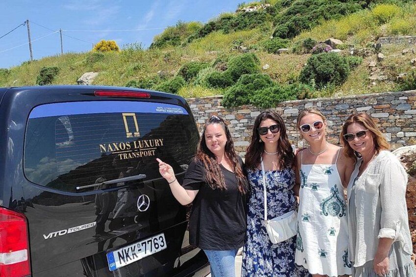 Un SUV negro con 'NAXOS LUXURY TRANSPORT' en la parte trasera, estacionado en una ladera con un muro de piedra y follaje al fondo. 2