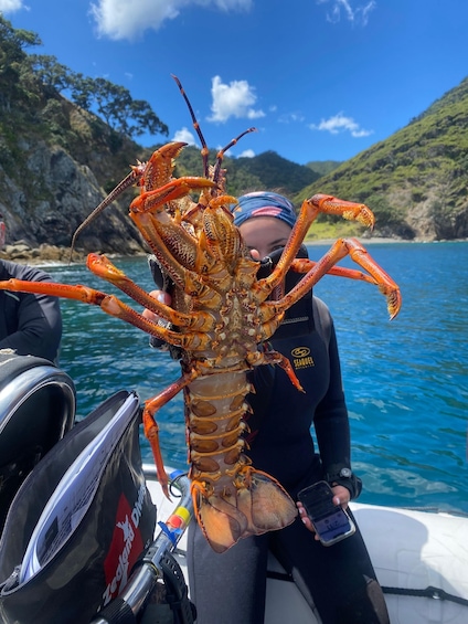 A person holding a large lobster on a boat with a clear blue sky and a mountainous landscape in the background. 2