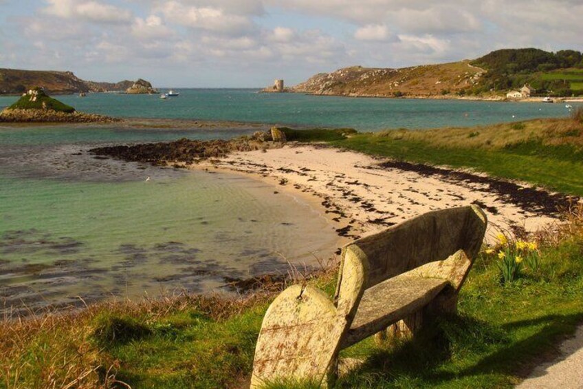A coastal scene with a bench, clear water, and distant hills. 4