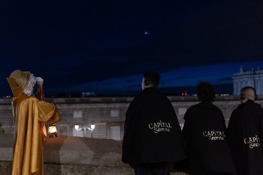 A statue of a historical figure, four people in black jackets with 'CAPITAL SECRET' text, and a large building in the background. 2
