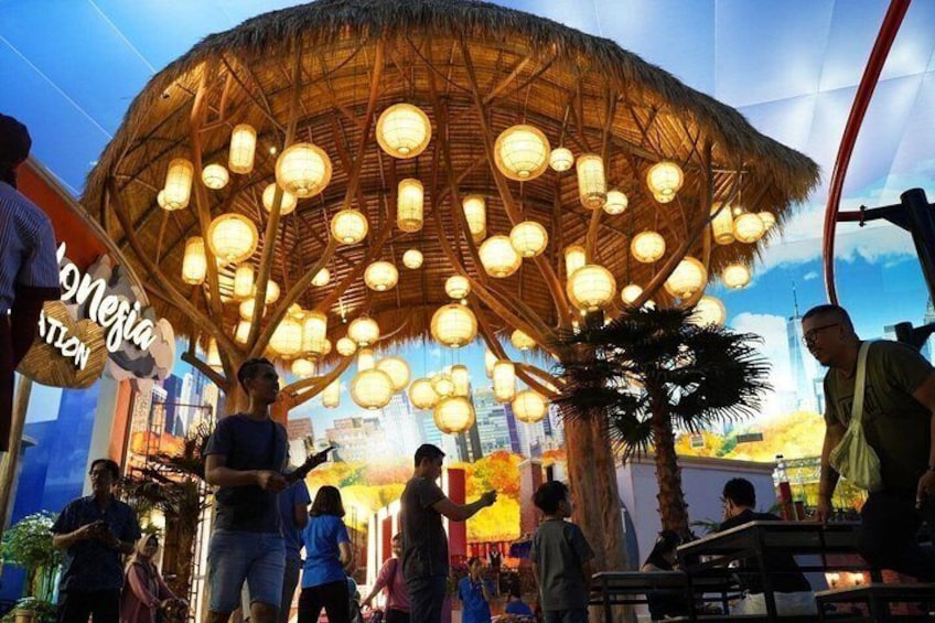 A large, open-air structure with a thatched roof and hanging lanterns, surrounded by palm trees and people. 1