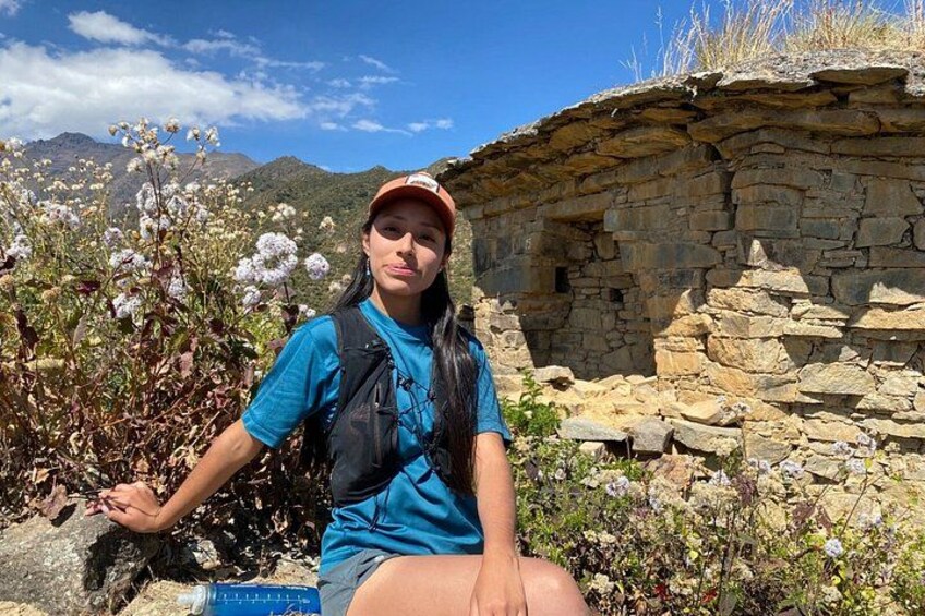 A person sitting on a rock with a stone structure and flowering plants in the background. 2