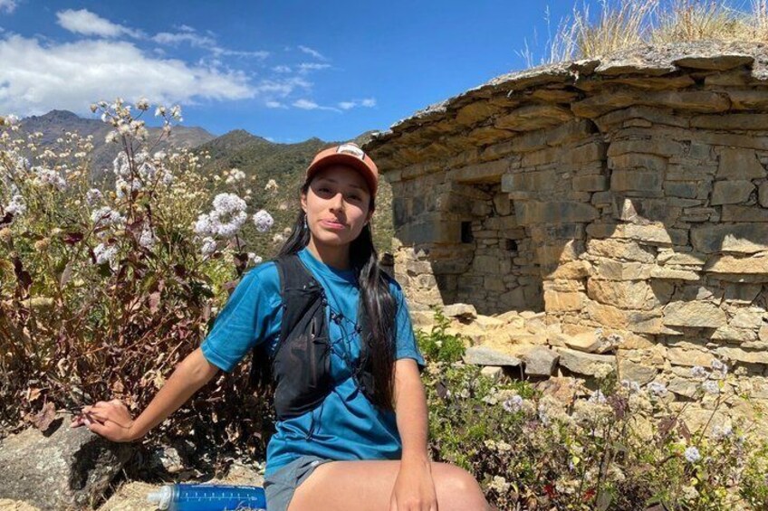 A person sitting by a stone structure with flowering plants and mountains in the background. 2
