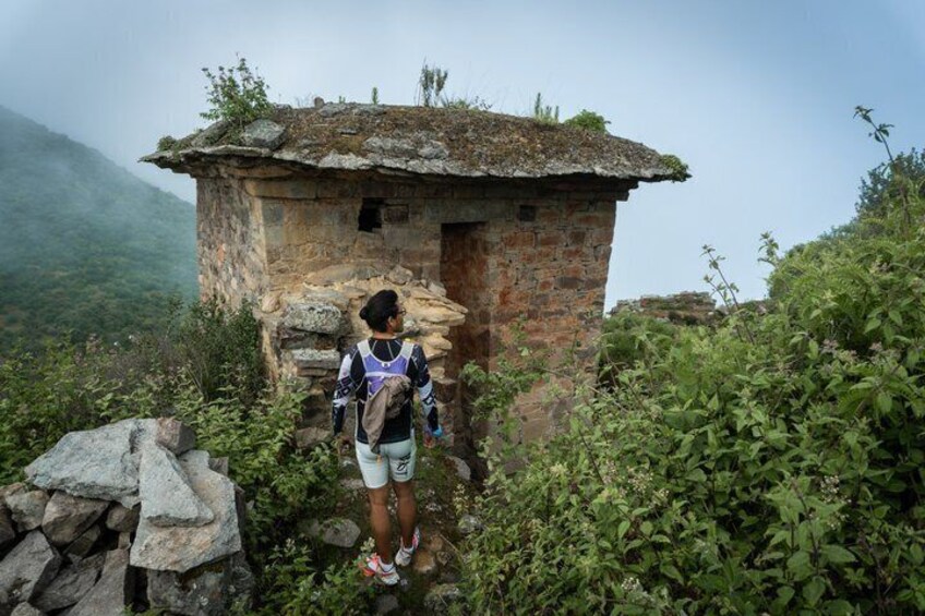 A hiker standing in front of a rustic stone structure with a thatched roof, surrounded by lush greenery and a mountainous backdrop. 1