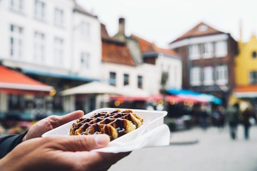 A hand holding a waffle with a paper liner, set against a blurred background of colorful buildings. 1