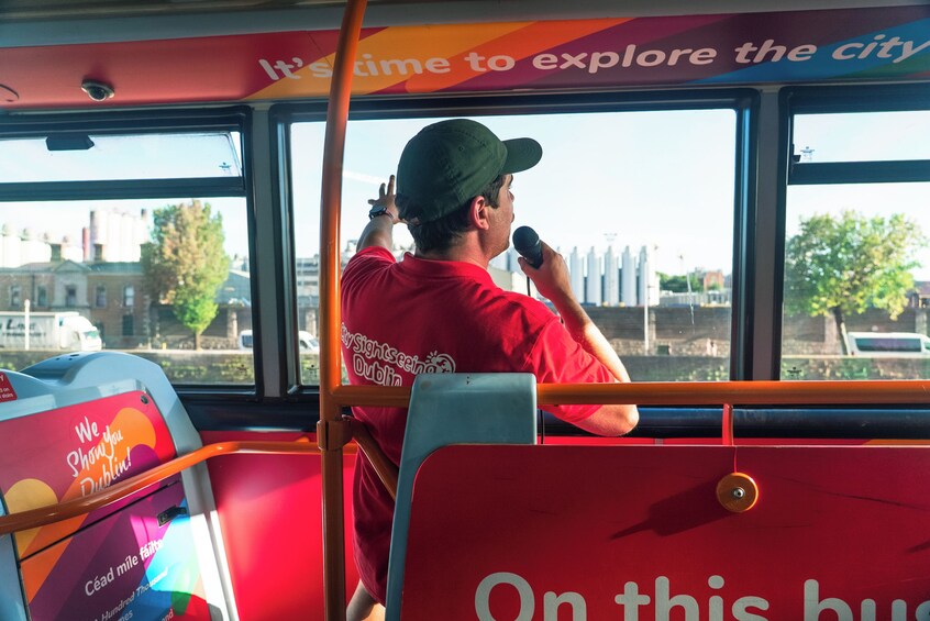 A person in a red shirt and cap, holding a microphone, sitting on a bus with a cityscape visible through the window. 5