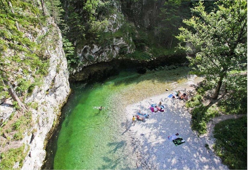 A rocky canyon with a clear green river, people sunbathing on the riverbank, and trees surrounding the area. 3