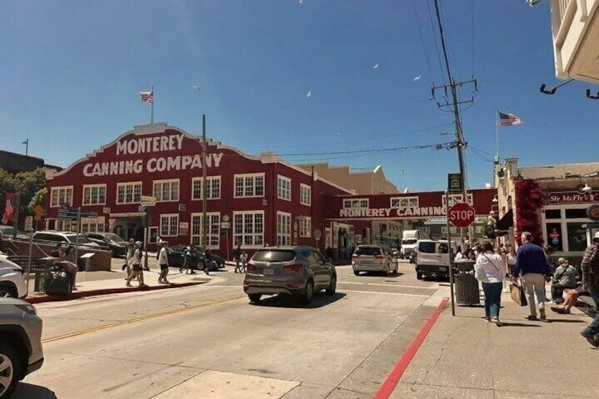 Un bâtiment rouge portant l’inscription « Monterey Canning Company », avec un panneau « STOP », des voitures dans la rue et des piétons. 1
