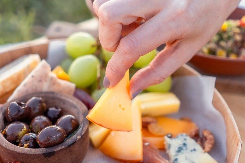 Una mano escogiendo una porción de queso de una tabla de madera con diversos quesos, uvas y aceitunas. 3