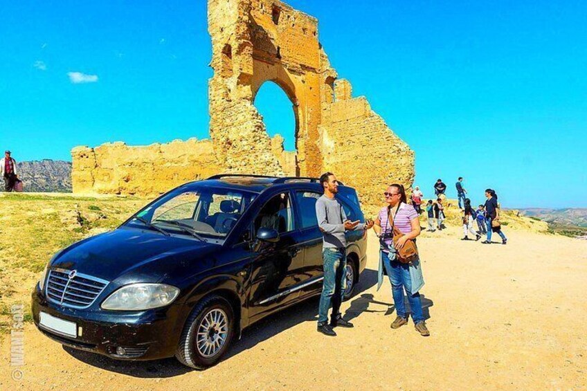 A black car parked in front of an ancient stone archway with tourists walking around. 1