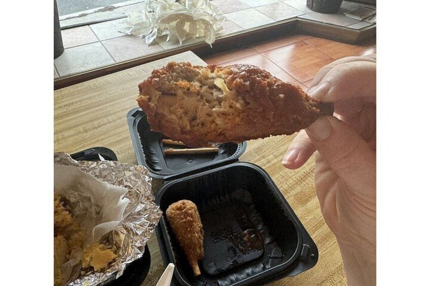 A hand holding a fried chicken wing, a small container with a bone, and a foil-wrapped food item on a table. 2