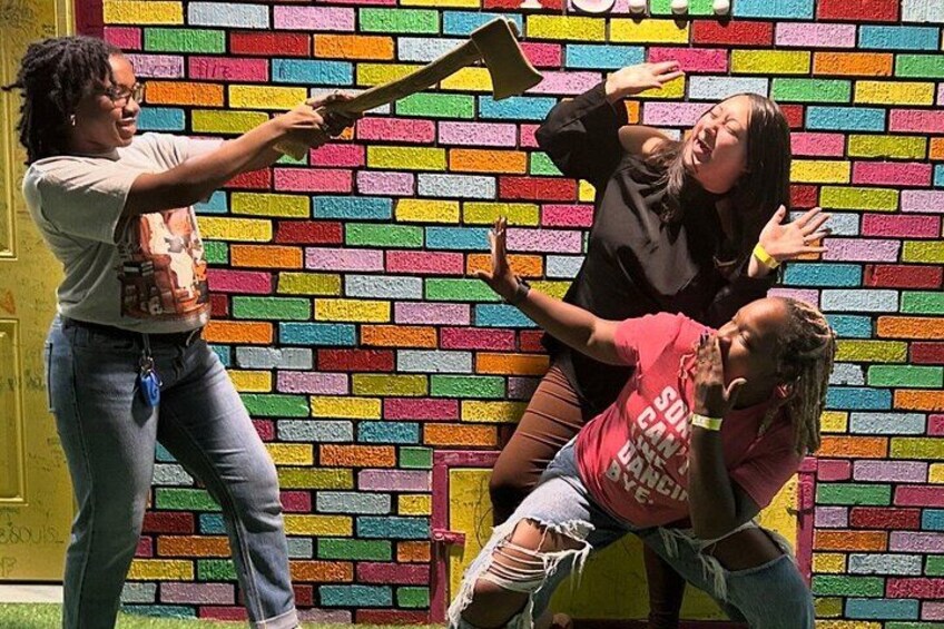 A person holding a bat, another person in a red shirt, and a third person in a black shirt, all in a room with a colorful brick wall. 3