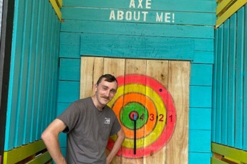 A man standing in front of a dartboard with the text 'AXE ABOUT ME!' on a teal-colored structure. 4