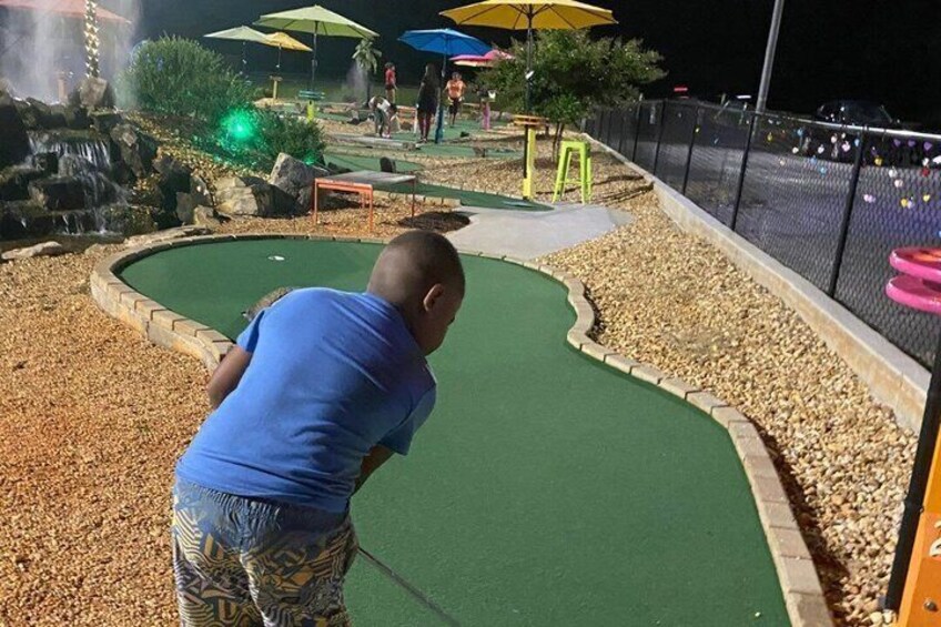 A miniature golf course at night with a green putting area, a water feature, and colorful umbrellas. 2