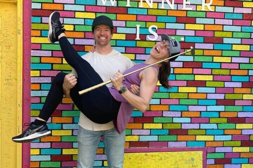 A person holding another against a colorful brick wall with the text 'WINNER IS'. 1