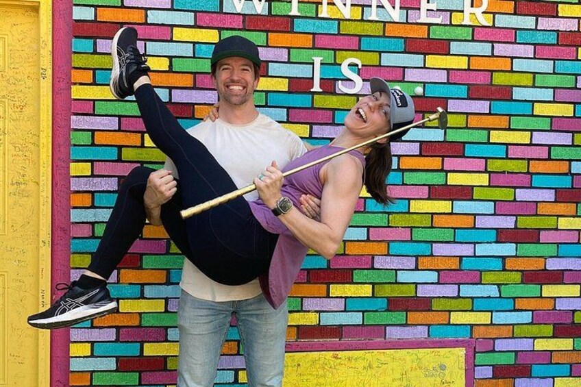 A person holding another person in a playful pose against a colorful brick wall with the text 'WINNER IS' partially visible. 1