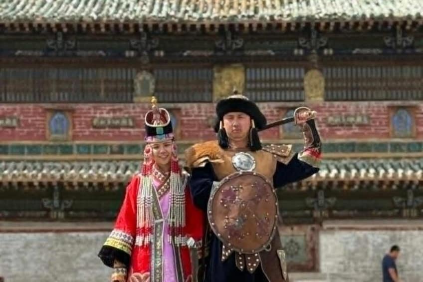Deux personnes, vêtues de costumes traditionnels, dont l’une tient un bouclier, se tiennent devant un bâtiment dont le toit est recouvert de tuiles. 1