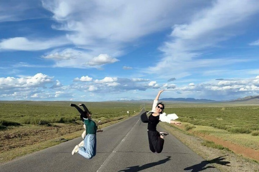 Two people jumping on a road with a flat landscape and distant mountains under a partly cloudy sky. 1