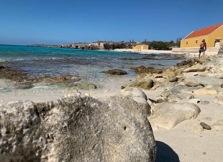 A rocky beach with clear turquoise water, a yellow building, and a person walking. 3