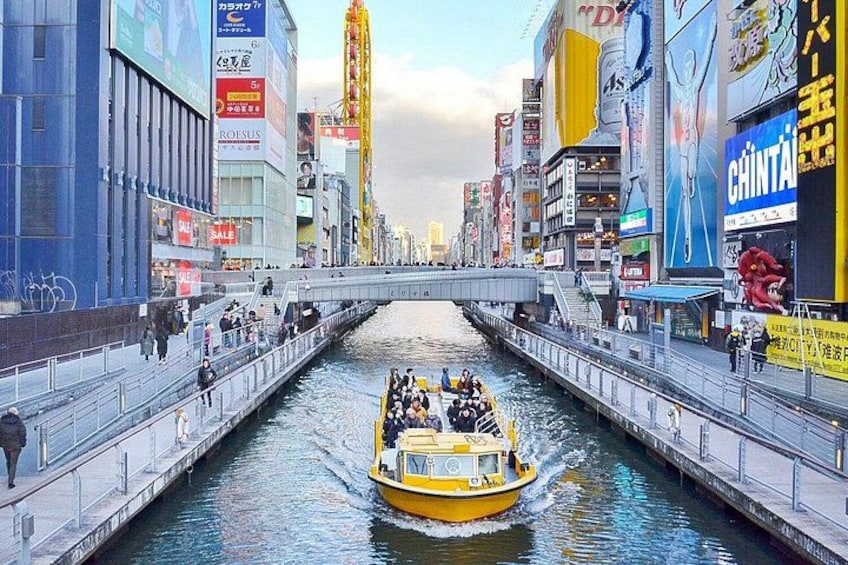 Un bateau jaune sur un canal, dans une ville où se dressent de hauts immeubles et où l’on peut voir de nombreux panneaux publicitaires. 1