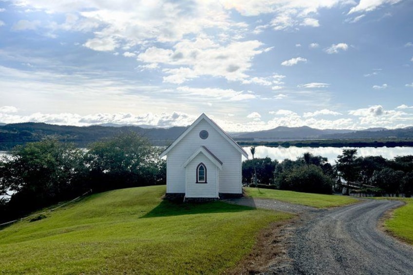 A white church with a steeple, surrounded by a grassy field and a dirt road, with mountains and a lake in the background. 3