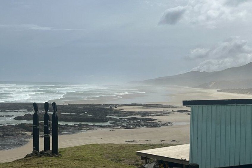 Een strand met rotsachtige delen, een houten constructie en verre bergen onder bewolkte luchten. 3