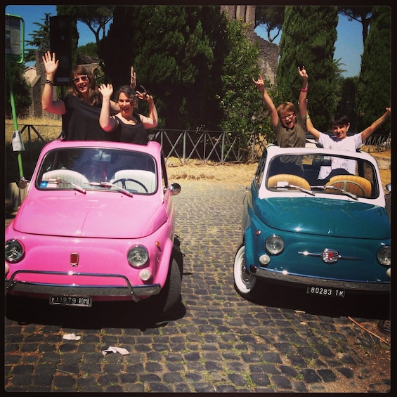 Two vintage cars, one pink and one teal, parked on a cobblestone street with people waving. 3