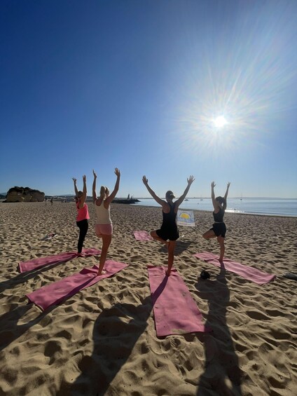 Four people performing yoga on pink mats on a sandy beach with the sun shining in the background. 2